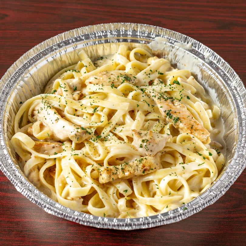 Creamy chicken fettuccine alfredo with parsley in a foil takeout container on a wooden table.