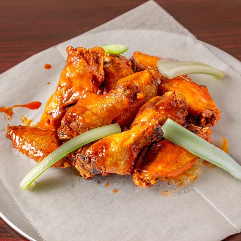 Topolino's buffalo wings, coated in orange sauce with celery sticks, on a white paper-lined plate.
